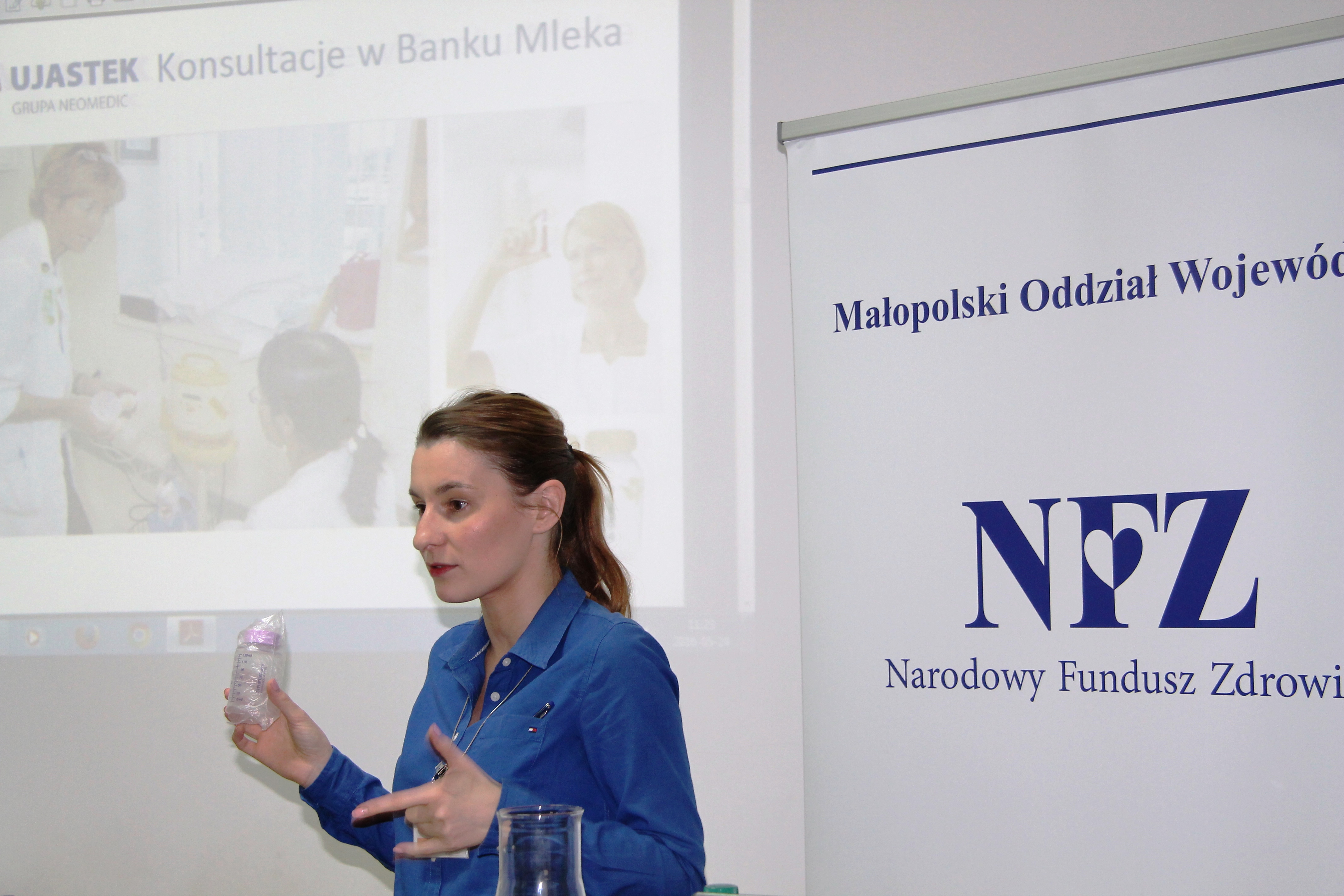 Dzień otwarty w Ujastku-kobieta z butelką do karmienia niemowląt soi na tle baneru z logo NFZ i prezentacji z napisem Ujastek. konsultacje w Banku Mleka 